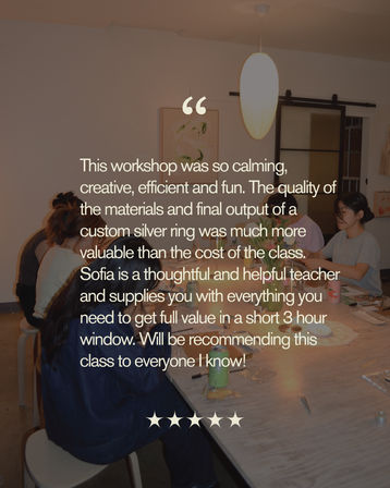 Cozy indoor jewelry-making workshop with participants seated around a wooden table covered in tools, supplies and drinks, warm pendant light overhead, and a large testimonial text overlay praising a calming, creative silver-ring class with a star-rating graphic.