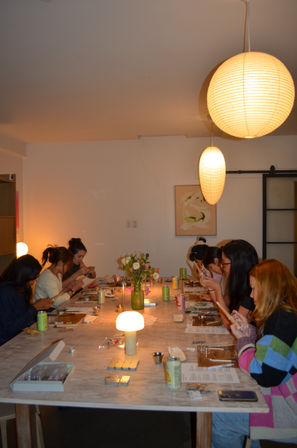 Participants at an evening DIY craft workshop seated around a long wooden table, lit by warm paper lanterns and table lamps.