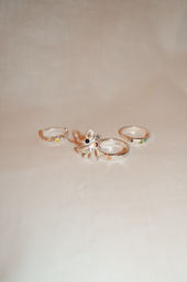 Studio flatlay of three silver rings on beige fabric: a floral statement ring with a dark center stone and two slim stacking bands with small green and yellow gemstone accents.