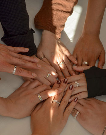 Circle of diverse hands on a white surface, each wearing silver gemstone rings and some with dark nail polish, bathed in warm natural sunlight.