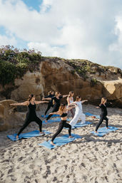 Seven women in activewear doing a beach yoga class on blue mats on a sandy shore beside sandstone cliffs and coastal plants under a partly cloudy sky