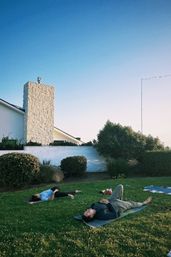Outdoor yoga session: two people lying on mats on a green residential lawn beside a white house with a stone chimney under a clear blue sky.