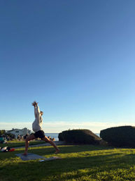 Person doing a high lunge yoga pose on a mat in a grassy seaside park at golden hour, with ocean and coastal homes in the background.