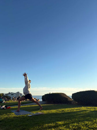 Person doing a high lunge yoga pose on a mat in a grassy seaside park at golden hour, with ocean and coastal homes in the background.