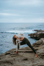 Barefoot woman practicing outdoor yoga in a deep side stretch on rocky coastal cliffs by the ocean under a cloudy sky