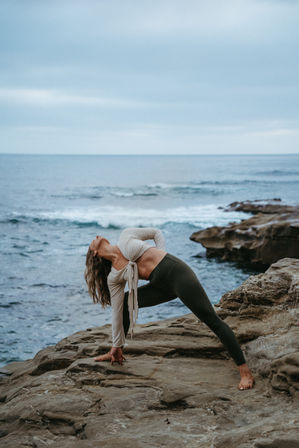 Barefoot woman practicing outdoor yoga in a deep side stretch on rocky coastal cliffs by the ocean under a cloudy sky