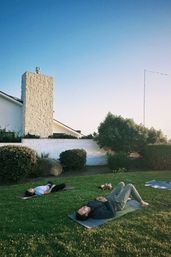 Two people lying on yoga mats on a green lawn in a suburban backyard at sunset, with a white stone chimney, trimmed shrubs and a clear blue sky