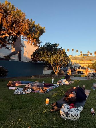Sun-soaked outdoor yoga class lying on mats on a grassy, palm-lined coastal park at golden hour, with a large wall mural and nearby houses in the background.