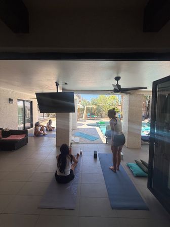 Poolside backyard yoga session on a covered patio — people on mats under a ceiling fan with a wall-mounted TV and a sunny pool and lounge area visible.