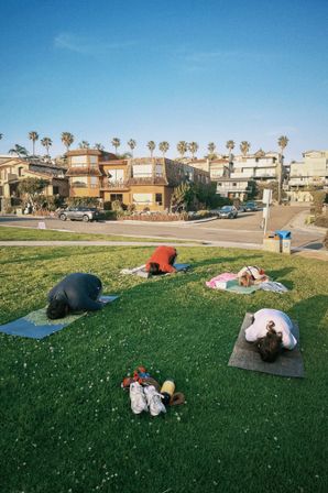 Four people folded into child's pose on yoga mats in a sunny beachside grassy park, palm trees and coastal houses lining the street behind them, with shoes and a water bottle in the foreground.