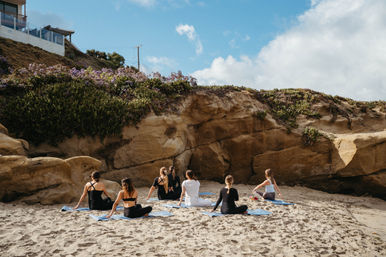 Group practicing beach yoga on blue mats in a sandy cove beneath rocky coastal cliffs topped with flowering greenery under a bright blue sky.