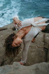 Woman performing a twisted side-plank yoga pose on coastal rocks above crashing ocean waves, wearing a knotted cream top and dark leggings.