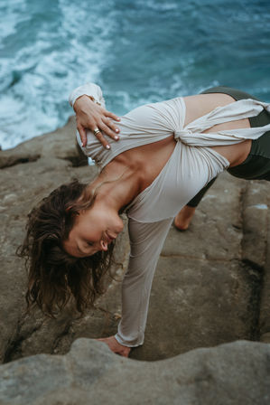 Woman performing a twisted side-plank yoga pose on coastal rocks above crashing ocean waves, wearing a knotted cream top and dark leggings.