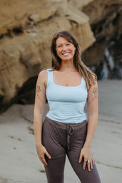 Smiling woman in a light blue tank top and brown drawstring leggings posing on a sandy beach in front of coastal rock cliffs, with matching floral tattoos on both upper arms and wearing rings and hoop earrings.