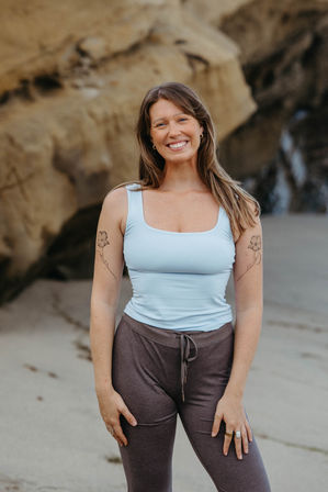 Smiling woman in a light blue tank top and brown drawstring leggings posing on a sandy beach in front of coastal rock cliffs, with matching floral tattoos on both upper arms and wearing rings and hoop earrings.