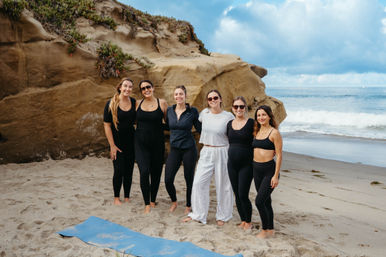 Six smiling women in athletic wear pose barefoot on a sandy beach beside sandstone cliffs and rolling ocean waves, a blue yoga mat lies in the foreground under a partly cloudy sky.
