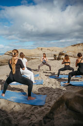 Group beach yoga class on blue mats practicing wide-legged squat poses on a sandy shore with sandstone rock formations and a dramatic cloudy blue sky