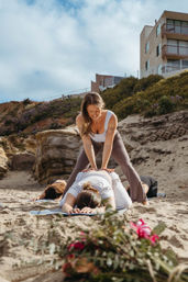Yoga instructor assisting a student in child's pose during a group beach yoga session on sandy coastal cliffs, mats and blurred flowers in the foreground under a blue cloudy sky