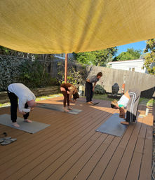 Outdoor yoga class on a sunny backyard wood deck — four people practicing forward bends on mats under a large yellow shade sail beside a decorative Buddha statue.