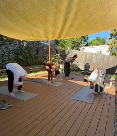 Outdoor yoga class on a sunny backyard wood deck — four people practicing forward bends on mats under a large yellow shade sail beside a decorative Buddha statue.