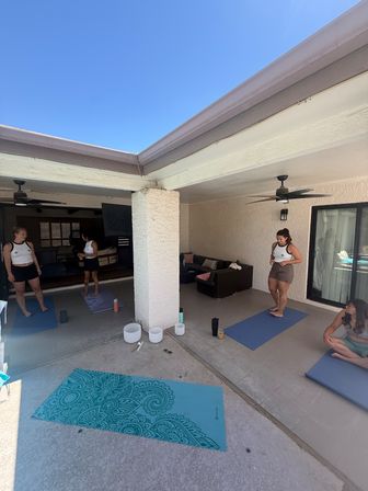 Sunny home patio yoga session with a small group on blue and teal mats under a covered terrace, ceiling fans, sliding glass doors, sofa seating and white crystal singing bowls