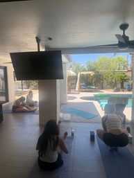 Outdoor yoga class on a shaded patio overlooking a sunny backyard pool with cabanas and palm trees.