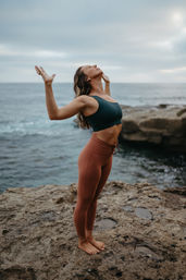 Fit woman practicing yoga on a rocky coastal cliff by the ocean, arms raised toward a cloudy sky in a peaceful wellness pose