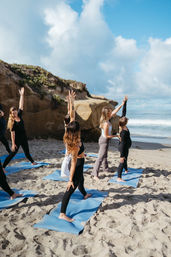Oceanfront beach yoga session: group on blue mats stretching arms on sandy shore beside sandstone cliffs under a bright, partly cloudy sky.