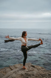 Woman practicing a serene standing yoga balance on a rocky coastal cliff overlooking the ocean under a cloudy sky.
