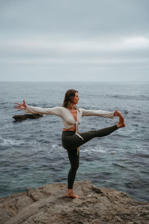 Woman practicing a serene standing yoga balance on a rocky coastal cliff overlooking the ocean under a cloudy sky.