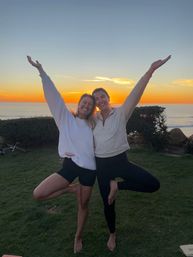 Two friends smiling and balancing in tree pose with arms raised on a grassy coastal bluff at sunset, orange sky and ocean horizon in the background.