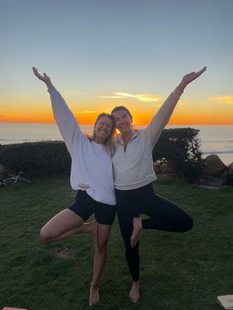 Two friends smiling and balancing in tree pose with arms raised on a grassy coastal bluff at sunset, orange sky and ocean horizon in the background.