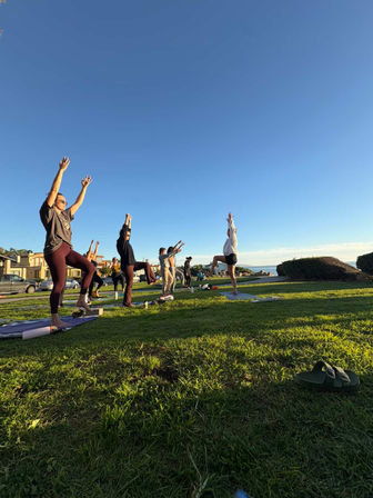 Group outdoor yoga class in a coastal grassy park at golden hour, people balancing in tree pose on mats with ocean and a clear blue sky behind them.