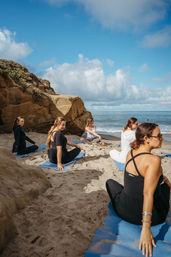 Group of women practicing seated yoga on blue mats on a sandy beach beside sandstone cliffs, with ocean waves and a bright blue sky.