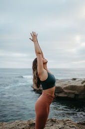 Person practicing yoga on a rocky coastline, arms raised toward a cloudy sky, teal sports bra and rust leggings with ocean waves in the background.