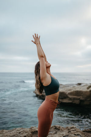 Person practicing yoga on a rocky coastline, arms raised toward a cloudy sky, teal sports bra and rust leggings with ocean waves in the background.
