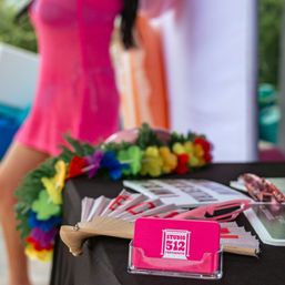Outdoor vendor table with bright pink business cards in a clear holder, colorful flower lei, wooden folding fan, sunglasses and photo prints on a black tablecloth, blurred person in a pink summer dress in the background.