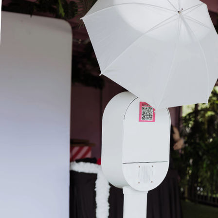 White outdoor photo booth kiosk with a large white umbrella and pink QR code sticker, positioned next to a blank event backdrop and garden greenery, ready for selfies