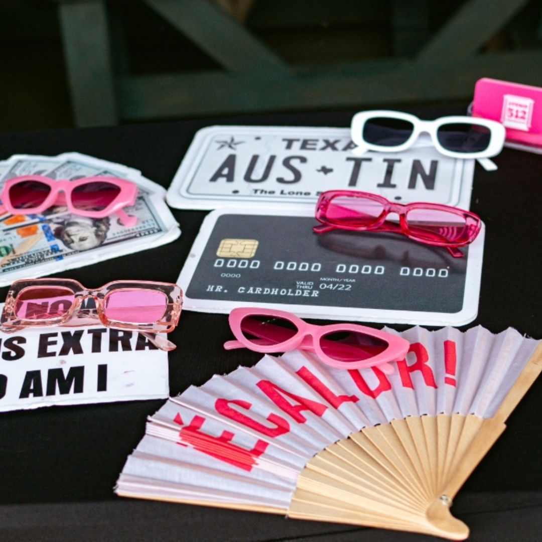 Playful tabletop display of multiple pink sunglasses and novelty props — an Austin, Texas license plate, a faux credit card, play money and a folding hand fan with red lettering.