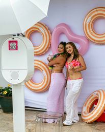 Two women in colorful bikinis pose with drinks at an outdoor poolside photo-booth backdrop decorated with a pink heart float and orange-striped inflatable rings, holding tropical cocktails under a white umbrella.