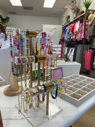 Colorful beaded bracelets and gold chain necklaces displayed on a three-tier acrylic and brass jewelry stand on a marble counter inside a bright clothing boutique with racks of patterned tops in the background.
