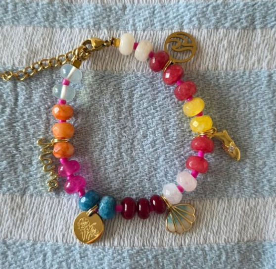 Vibrant multicolor beaded charm bracelet with glossy stone beads and gold-tone charms (shell, floral disc, scripted charm) laid on a blue-and-white striped towel — playful summer beach accessory
