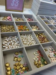 Close-up of a divided jewelry display tray filled with gold and enamel charms—seashells, starfish, floral pieces—pearl beads and small metal findings, $3 sign visible in background.
