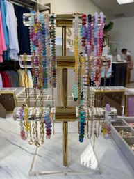 Colorful beaded bracelets and gold chain necklaces displayed on a three-tier acrylic and brass jewelry stand on a marble counter in a boutique shop.