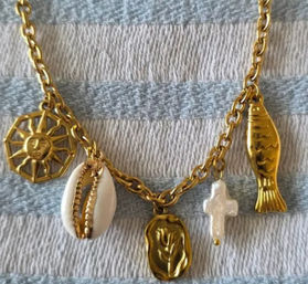 Gold charm necklace on blue-and-white striped fabric featuring a sun medallion, cowrie shell, wax-seal floral pendant, irregular freshwater pearl cross, and gold fish charm