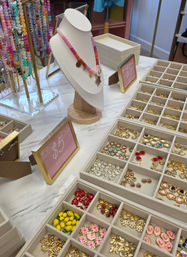 Playful boutique jewelry display on a marble table: pink ombré beaded necklace on a white bust, racks of colorful beaded chains, and compartment trays filled with gold and enamel charms next to a $5 sale sign