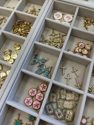 Close-up of a gray jewelry organizer tray filled with gold and enamel charms: iridescent blue butterfly pendants, sparkling crystal cross charms, pink sand-dollar discs, palm-tree rectangular tags and sunburst studs.