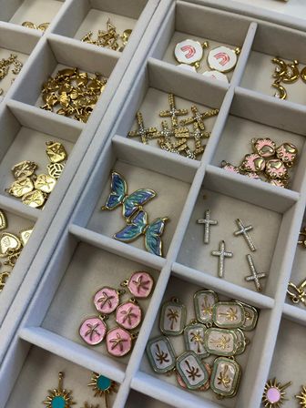 Close-up of a gray jewelry organizer tray filled with gold and enamel charms: iridescent blue butterfly pendants, sparkling crystal cross charms, pink sand-dollar discs, palm-tree rectangular tags and sunburst studs.