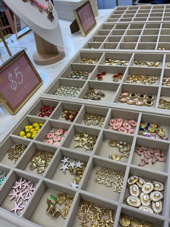 Organized display tray of small gold and colorful enamel jewelry charms and pendants in cubby compartments on a white marble counter with a pink $5 price sign
