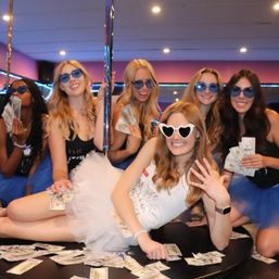 Bachelorette party friends in blue tutus and sunglasses at an indoor pole-party venue, bride-to-be in a white tutu and heart-shaped shades surrounded by dollar bills.